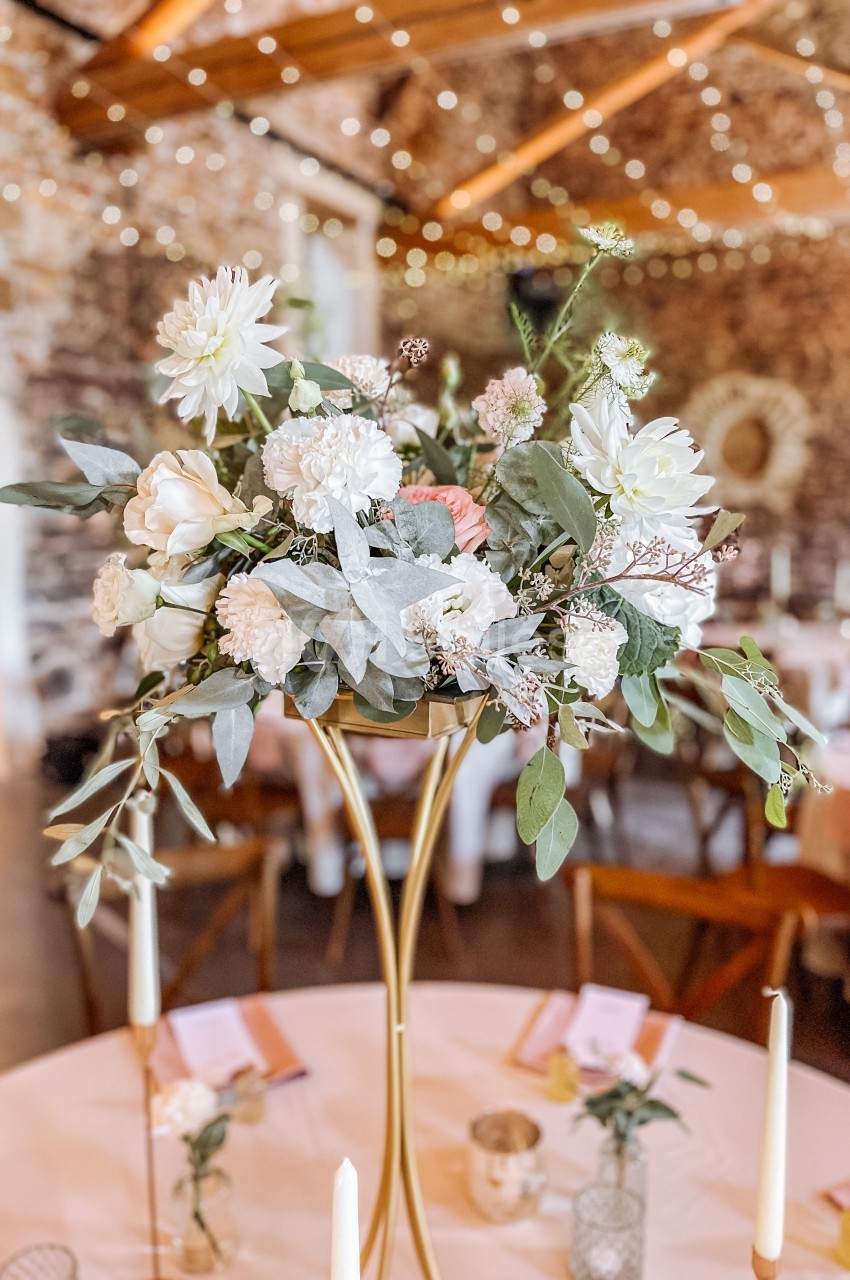 Centre de table floral avec des fleurs blanches et feuillage, posé sur un support doré dans une salle décorée de guirlandes…