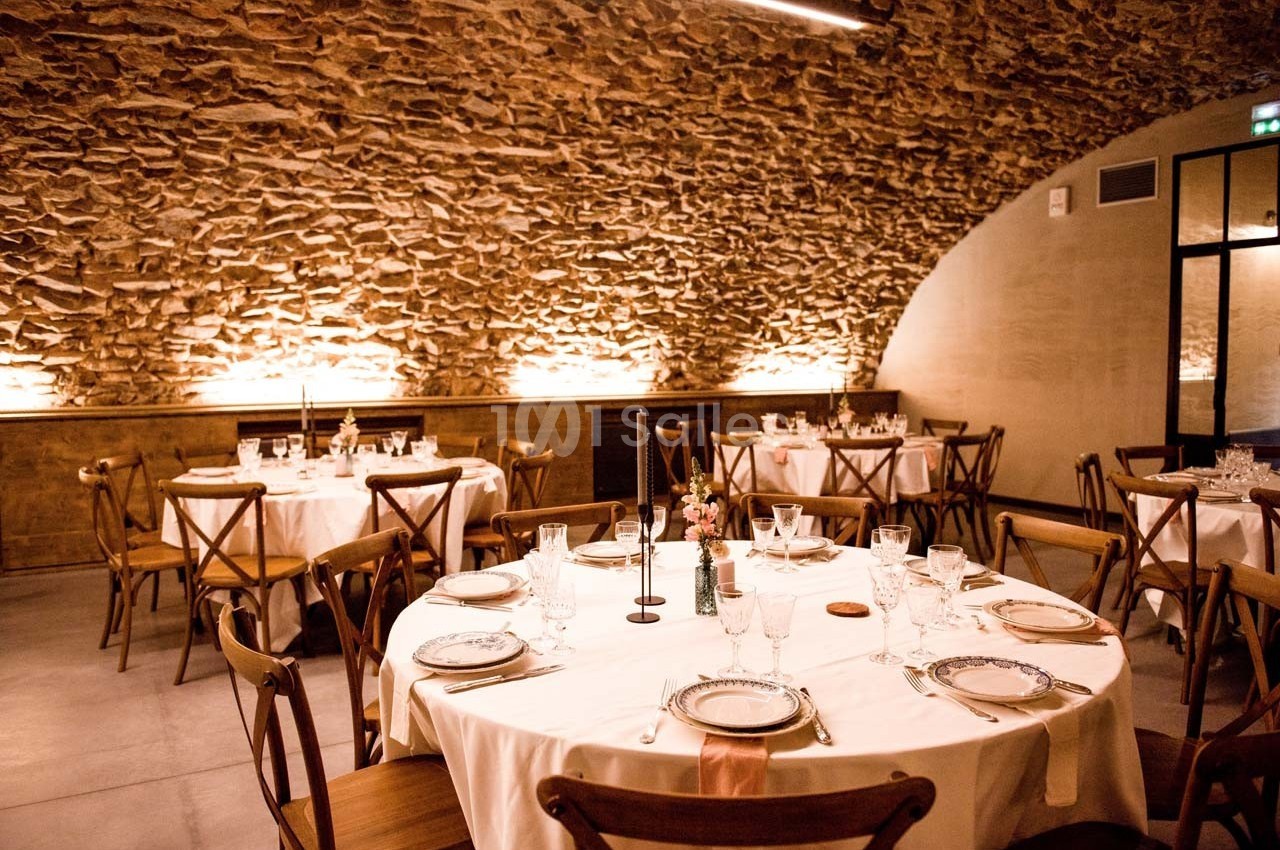 Salle voûtée en pierre avec tables rondes dressées pour un repas, décorée de bougies et vaisselle élégante.