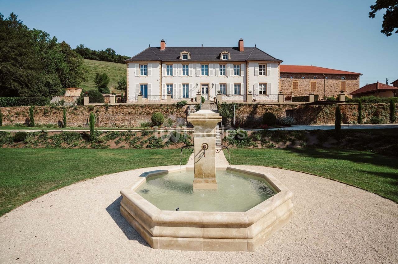 Fontaine octogonale en pierre au premier plan, devant une grande maison de campagne avec volets bleus et jardin aménagé.