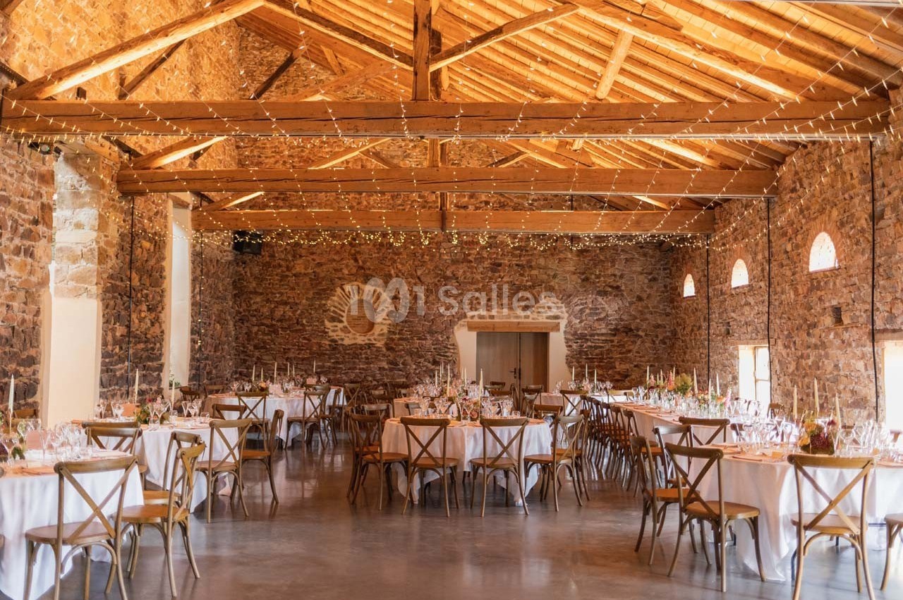 Salle en pierre avec poutres apparentes, tables rondes dressées pour un événement, éclairage chaleureux et guirlandes…