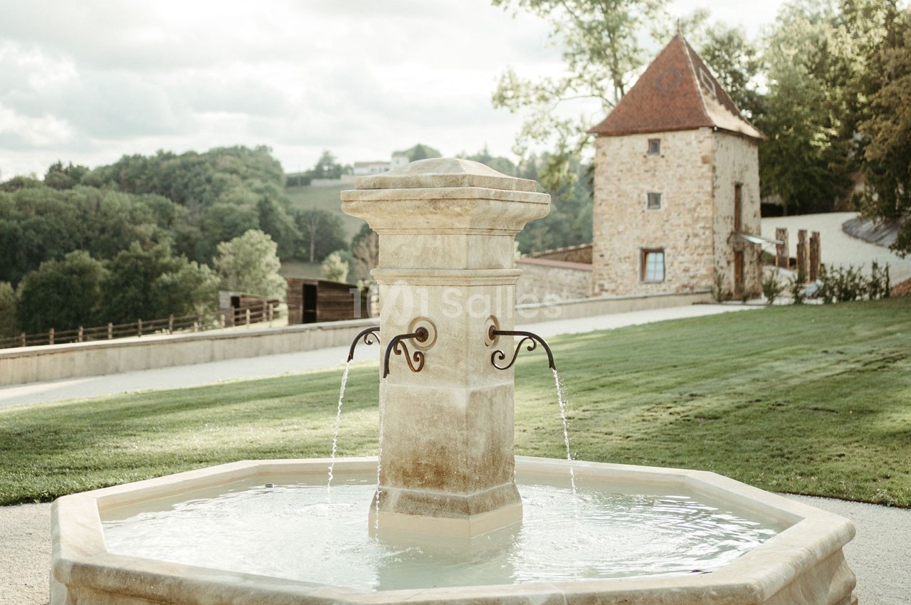 Fontaine octogonale en pierre avec jets d'eau, située dans un jardin verdoyant près d'une bâtisse en pierre.