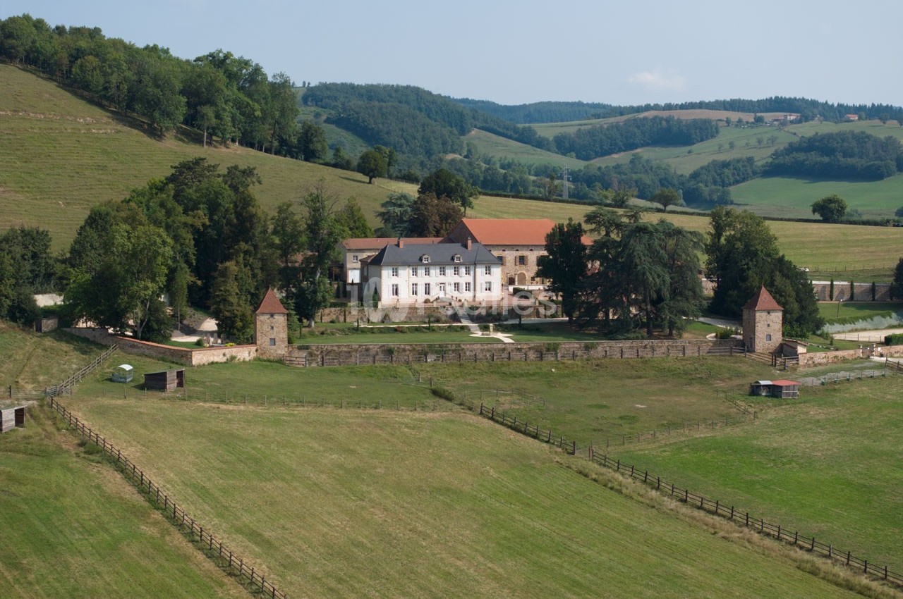 Vue d'un domaine rural avec une grande maison entourée de verdure et de collines boisées.