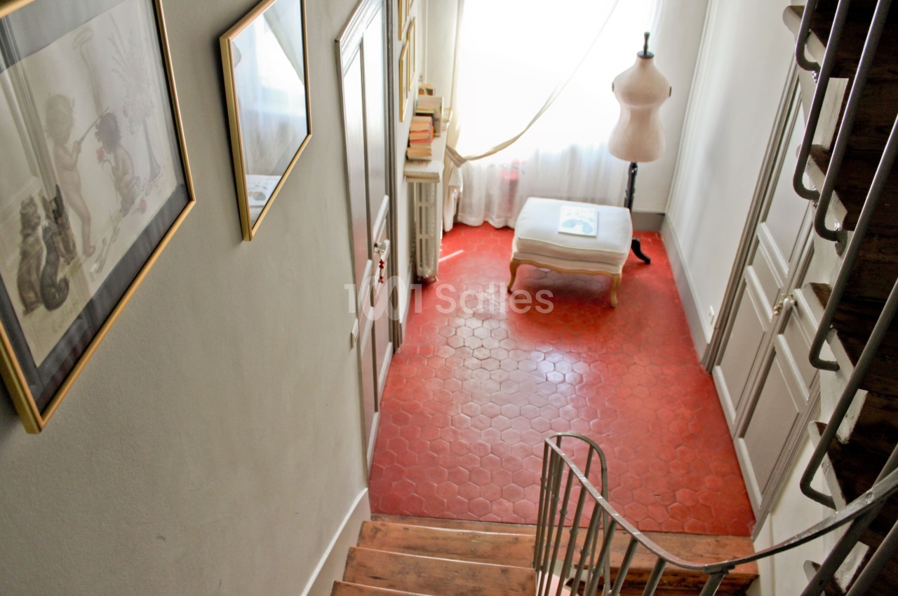 Vue d'un escalier en bois menant à un couloir avec sol en tomettes rouges, un mannequin et une fenêtre lumineuse.