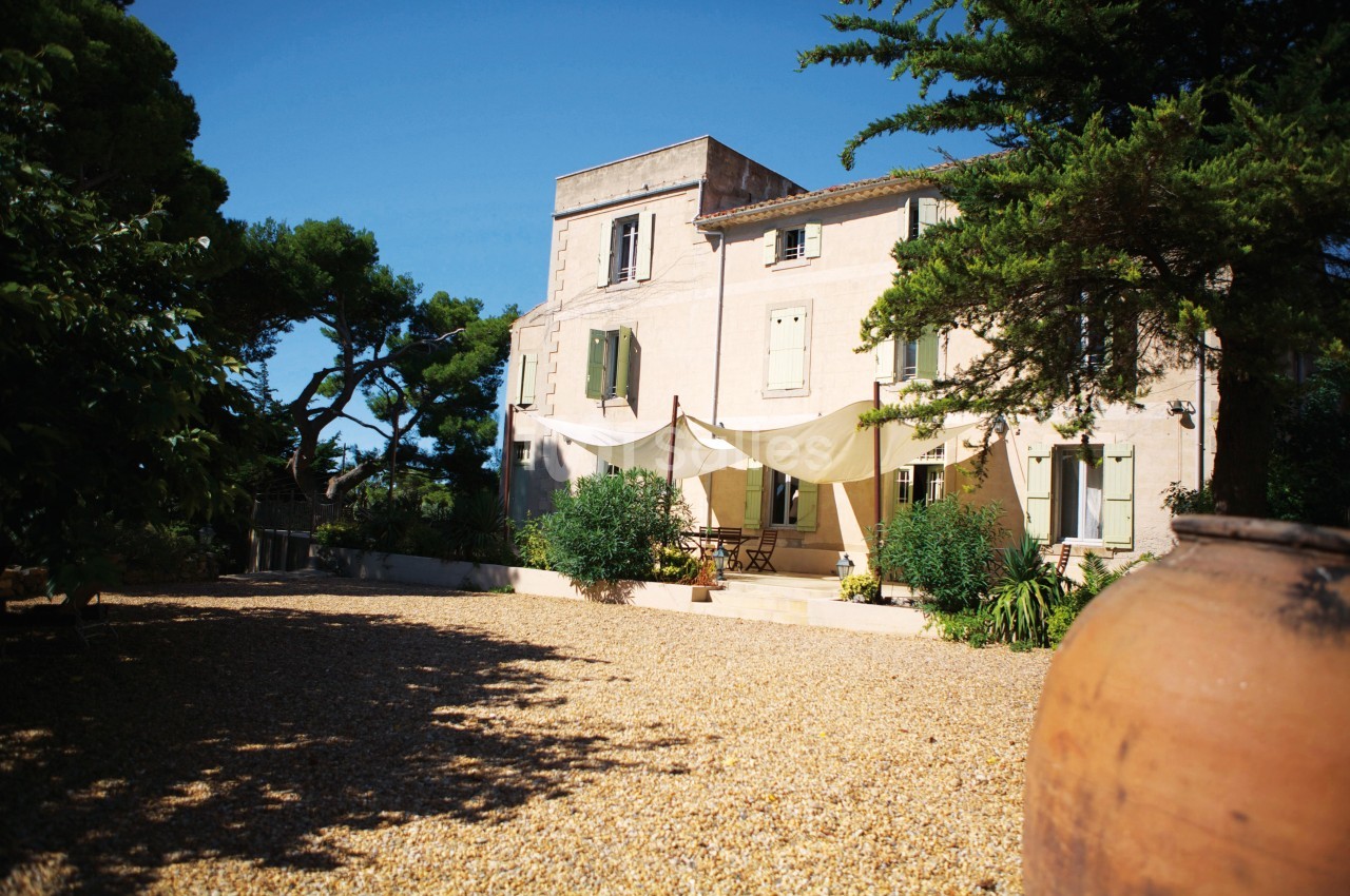 Grande maison en pierre avec volets verts, terrasse ombragée par des toiles, entourée de végétation et d'une cour…