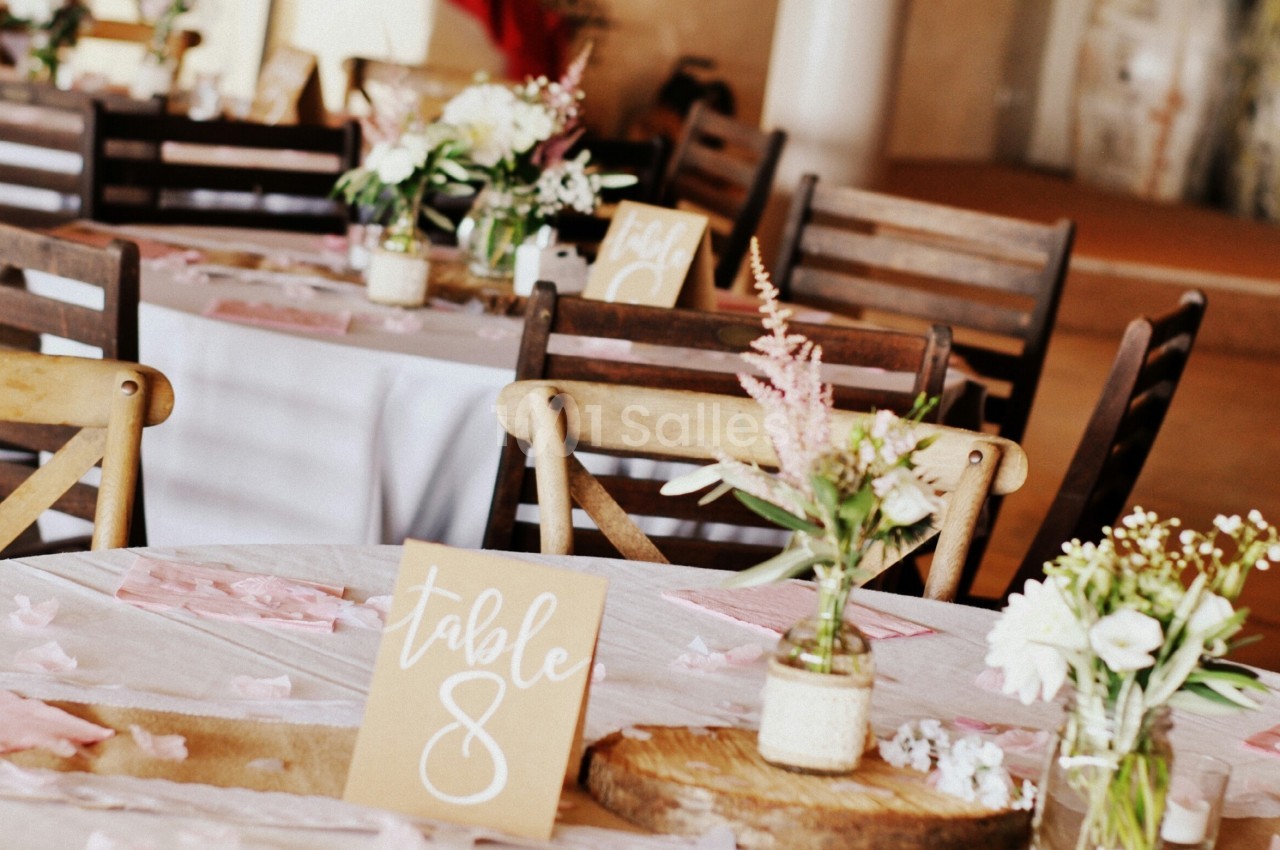 Salle décorée pour un événement avec des tables dressées, des fleurs et des panneaux indiquant les numéros des tables.