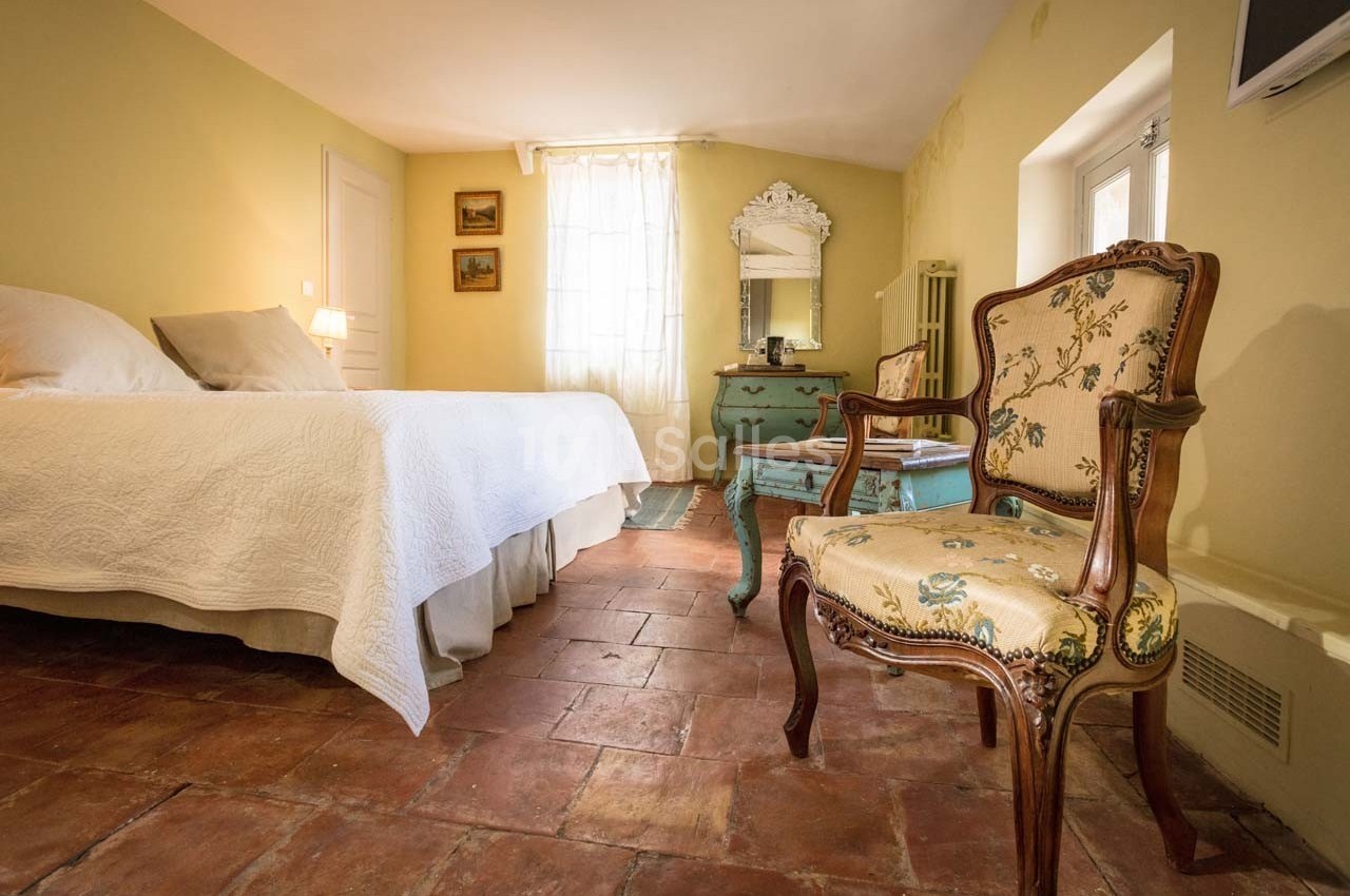 Chambre lumineuse avec lit double, fauteuil ancien, commode avec miroir et sol en carreaux de terre cuite.