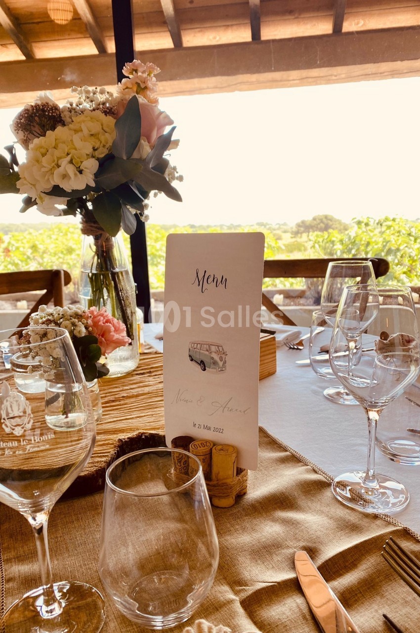 Table élégamment dressée avec fleurs, verres et menu, vue sur un paysage extérieur ensoleillé à travers une baie vitrée.