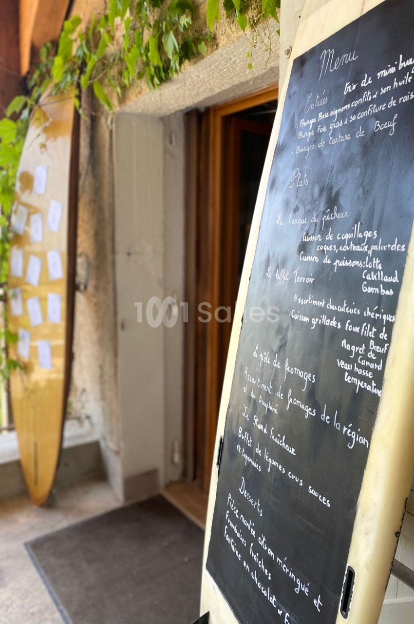 Ardoise avec menu écrit à la craie, posée devant l'entrée d'un restaurant avec une planche de surf en arrière-plan.