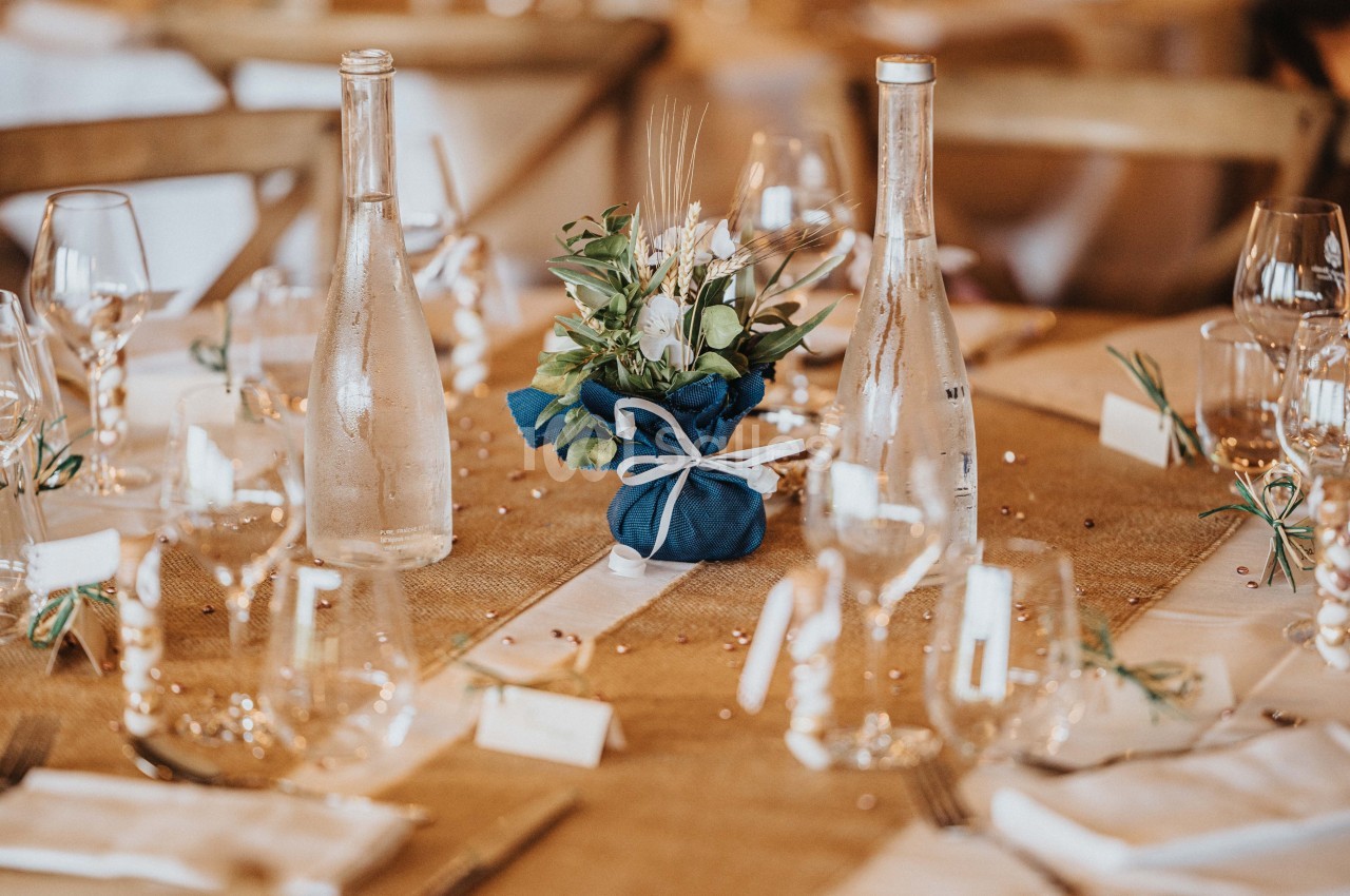 Table décorée avec nappes en jute, bouteilles en verre, verres, et un centre de table floral dans un tissu bleu.
