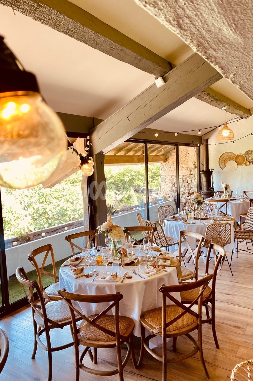Salle de réception lumineuse avec tables rondes dressées, chaises en bois et vue sur un extérieur verdoyant.