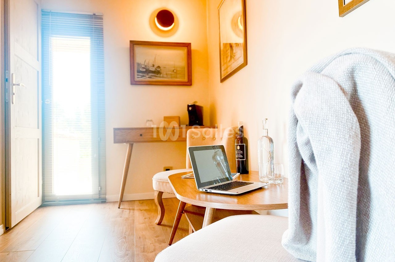 Un salon lumineux avec une table basse, un ordinateur portable, une bouteille d'eau et un plaid posé sur une chaise.