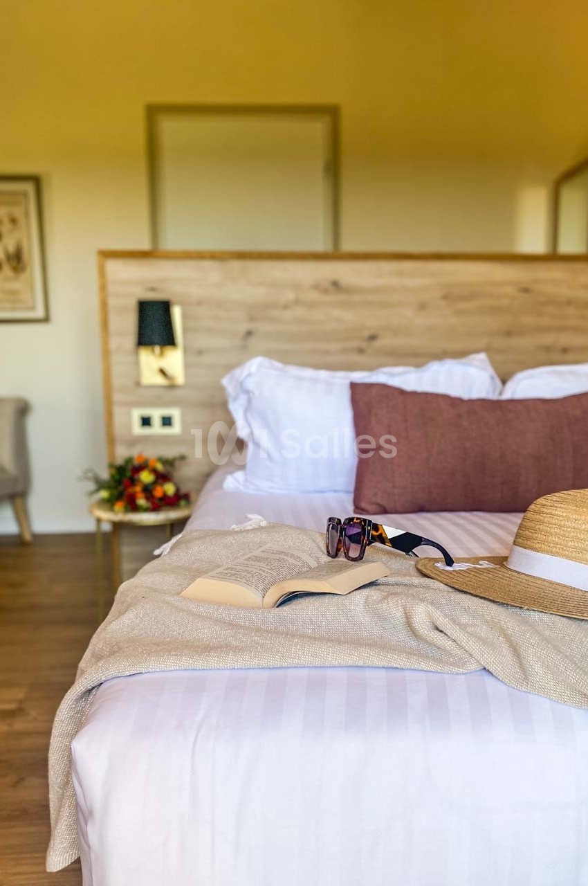 Lit double avec coussins, chapeau, lunettes de soleil et livre posé dessus, dans une chambre lumineuse au décor chaleureux.