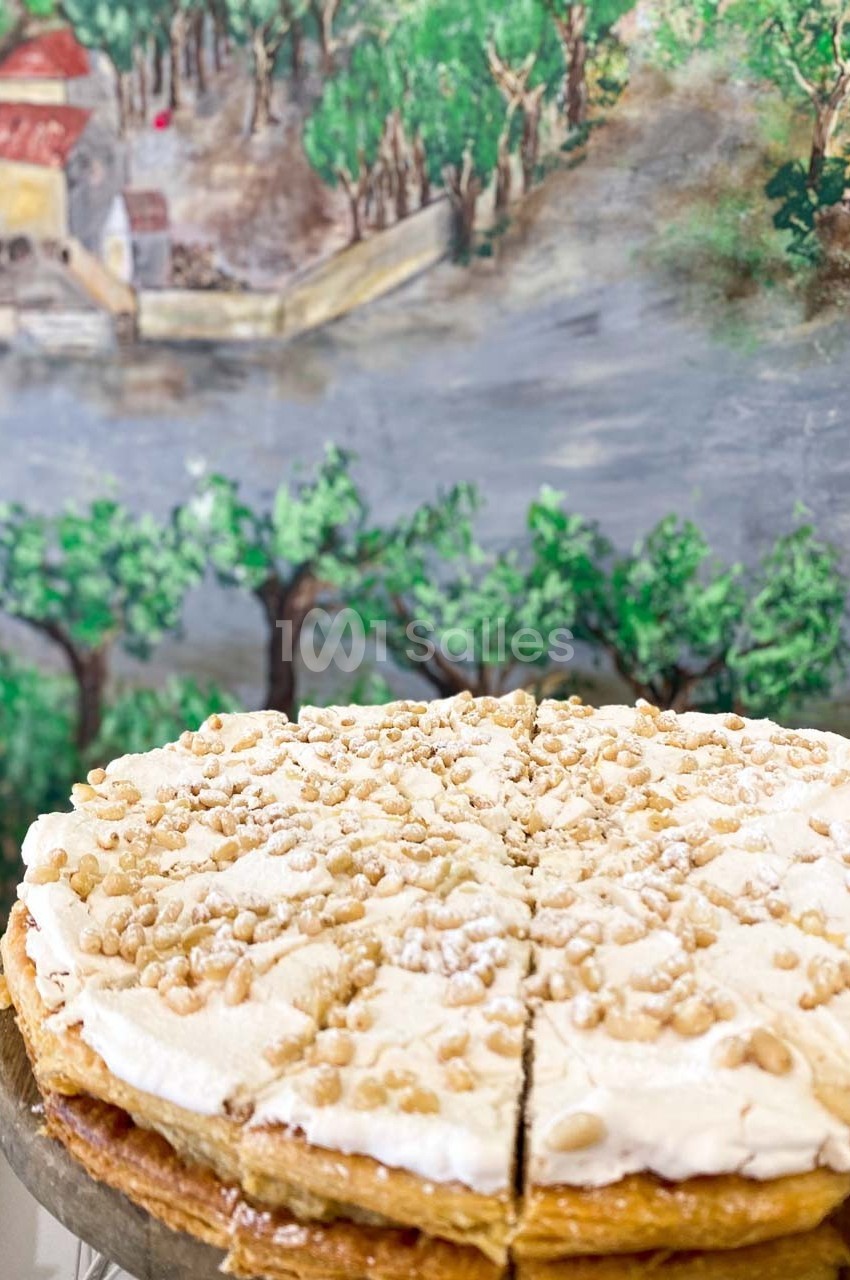 Tarte garnie de crème et saupoudrée de pignons de pin, posée devant une fresque représentant un paysage rural.