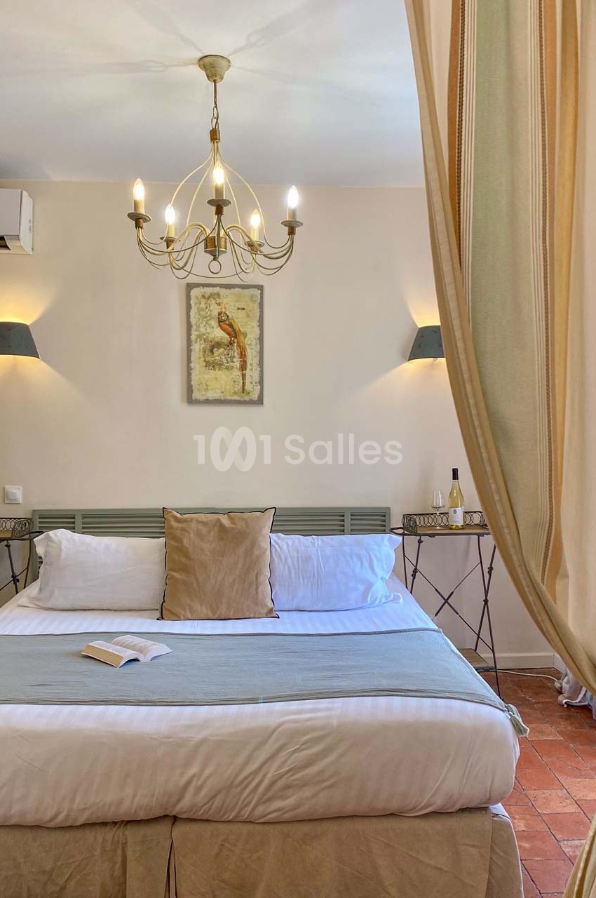 Lit double avec coussins, draps clairs, tables de chevet, rideaux ouverts, lustre et tableau mural dans une chambre…