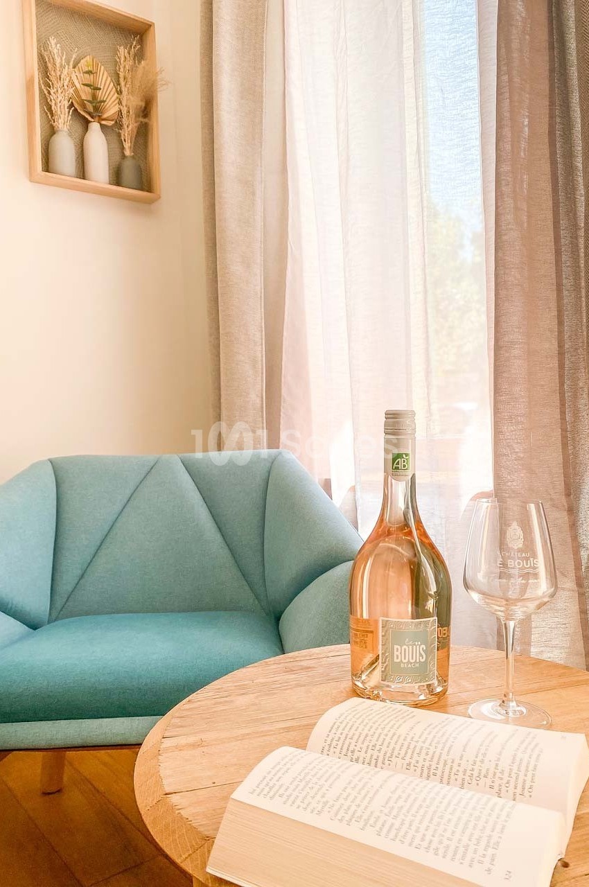 Une bouteille de vin rosé, un verre, un livre ouvert sur une table ronde près d'un fauteuil bleu et d'une fenêtre.
