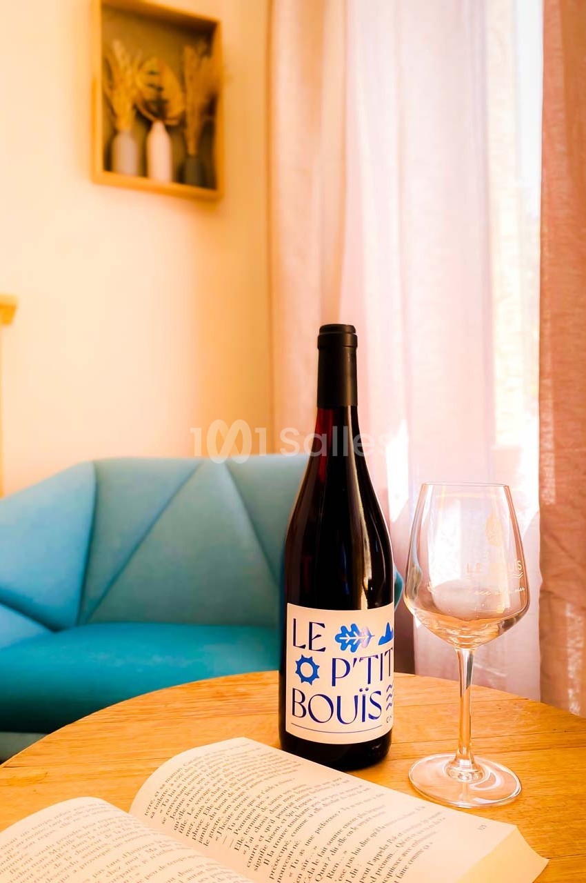Bouteille de vin rouge et verre vide posés sur une table en bois, avec un canapé bleu et une fenêtre en arrière-plan.