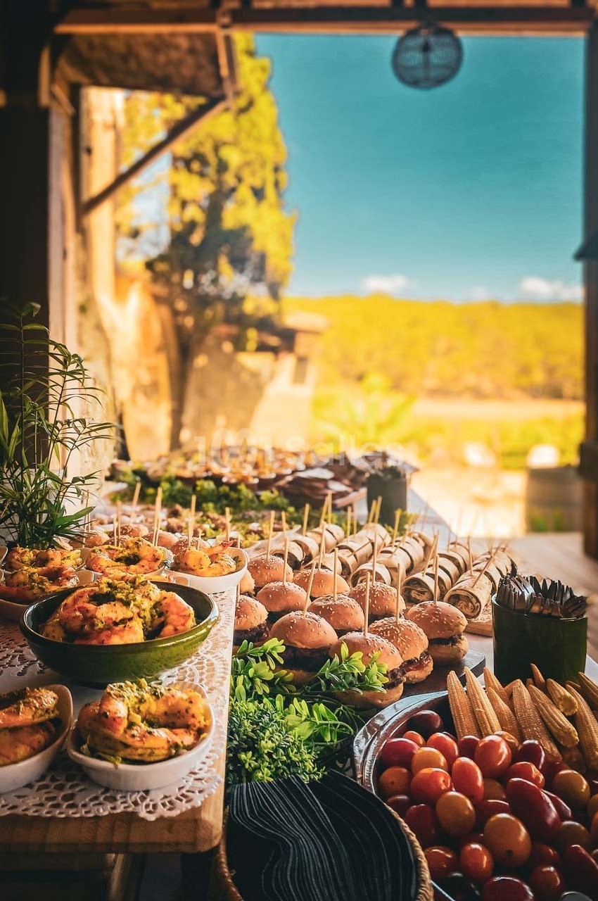 Buffet en extérieur avec divers plats, dont mini-burgers, légumes frais, épis de maïs et salades, sous une lumière naturelle.