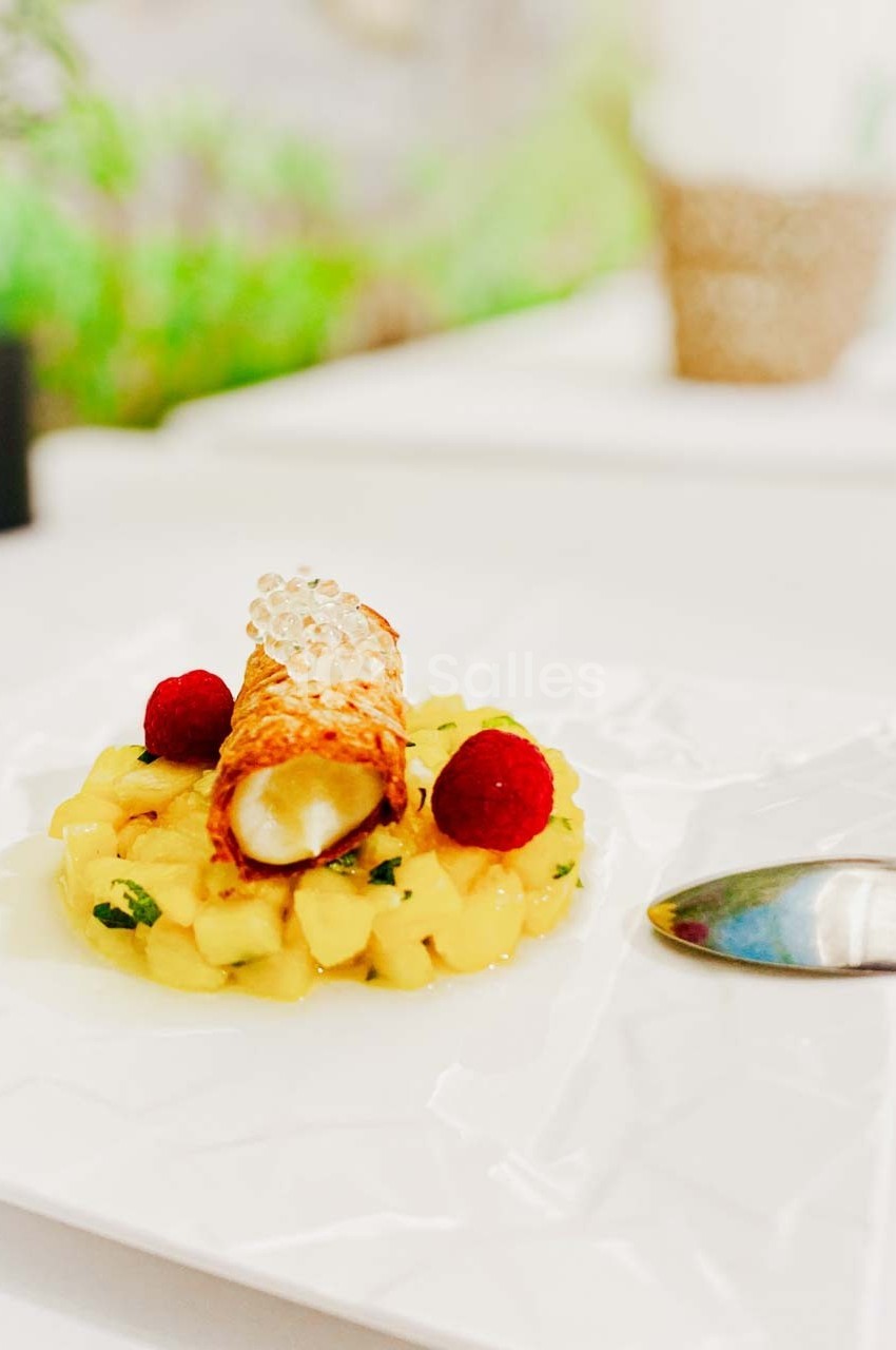 Dessert composé d'un cannolo garni, entouré de dés de fruits frais, décoré de framboises et de perles translucides.