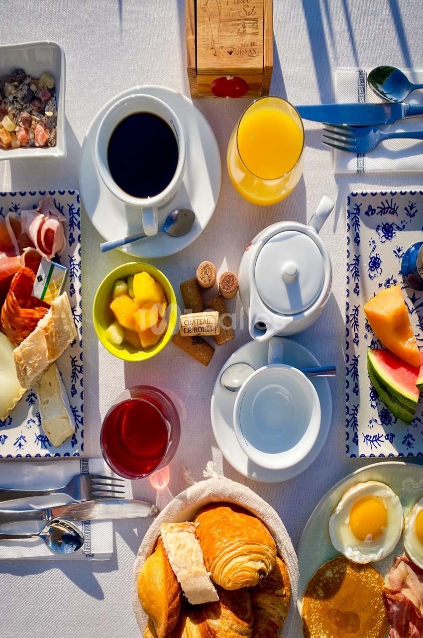 Petit-déjeuner varié avec viennoiseries, fruits, œufs, charcuterie, fromages, café, jus d'orange et thé sur une table…