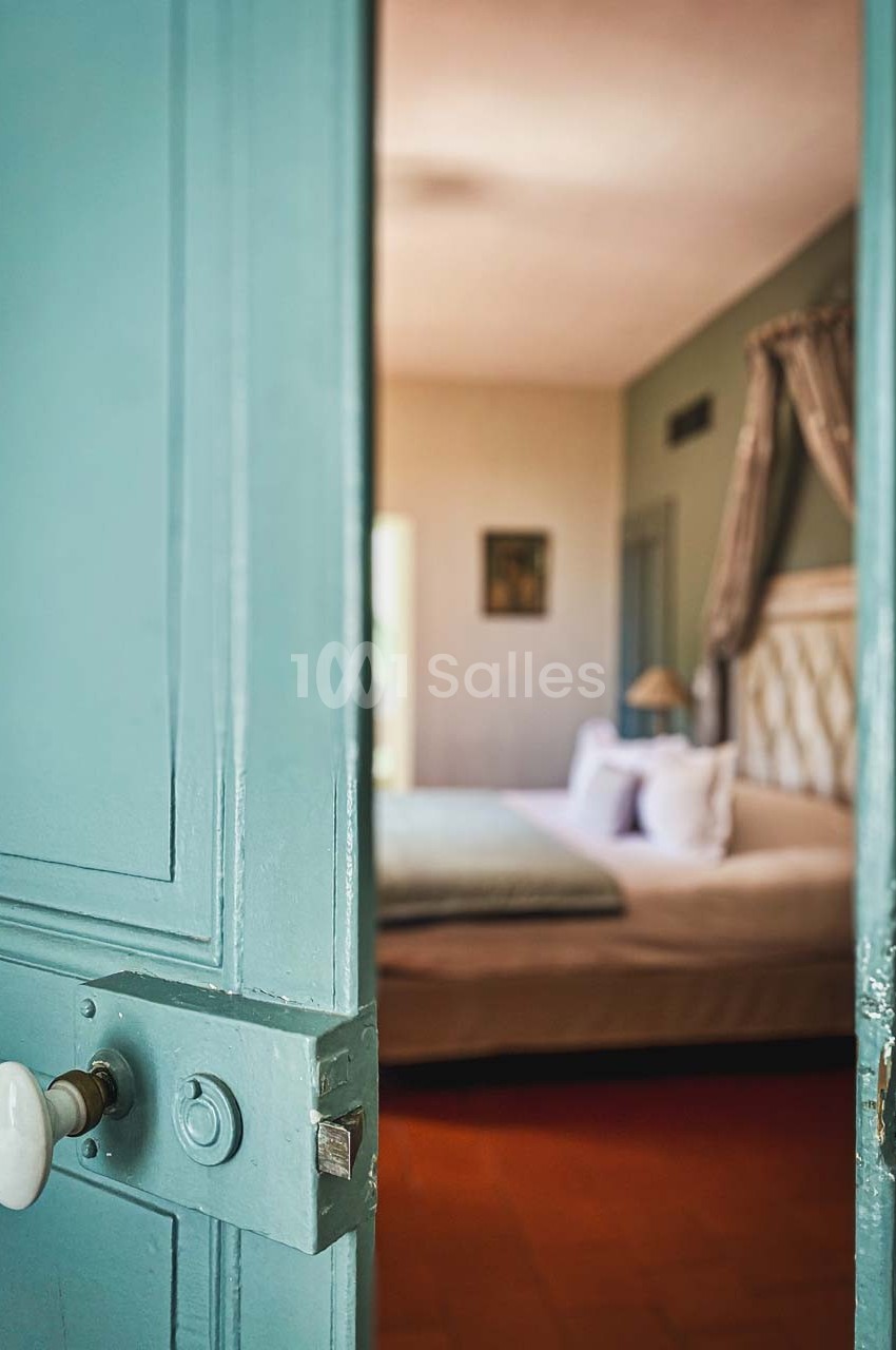 Porte bleue entrouverte donnant sur une chambre lumineuse avec un lit double et des draperies.