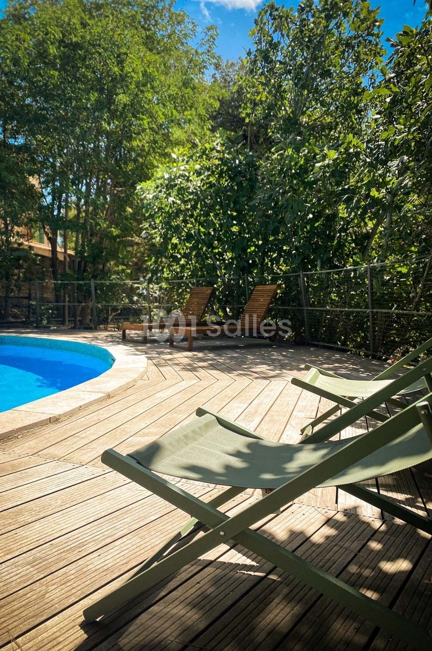 Chaises longues vertes sur une terrasse en bois près d'une piscine, entourée de végétation dense sous un ciel bleu.