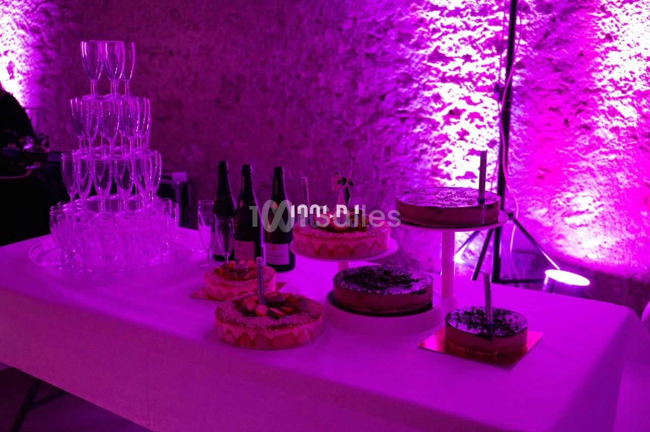 Table décorée avec des gâteaux, des bouteilles et une pyramide de verres, éclairée par une lumière violette.