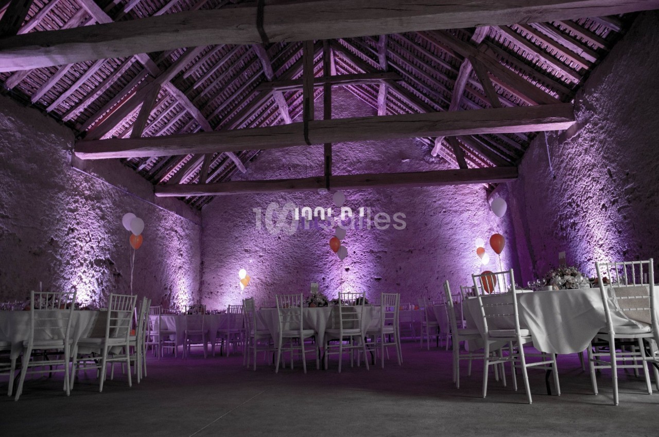 Salle en pierre avec charpente apparente, éclairée en violet, décorée de ballons et tables dressées pour un événement.