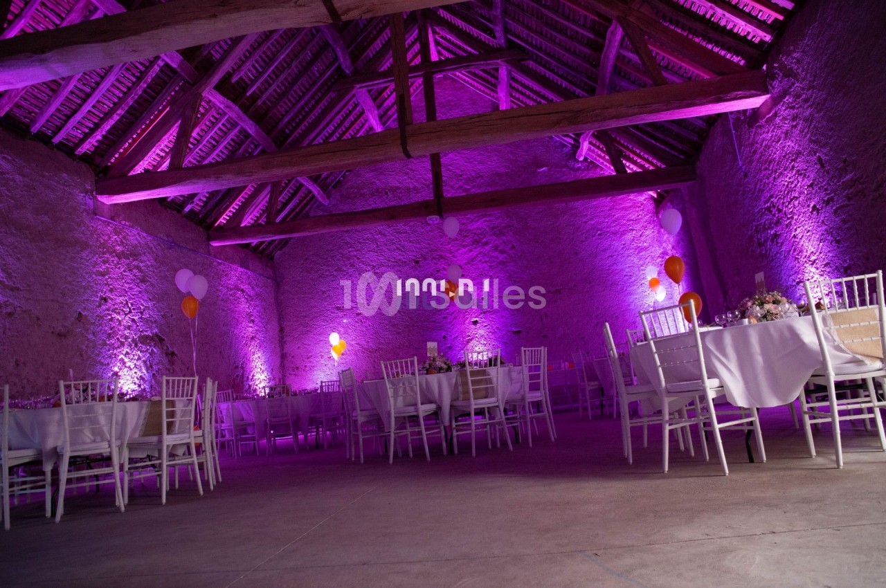 Salle de réception en pierre avec poutres apparentes, éclairée par des lumières violettes, tables et chaises blanches…