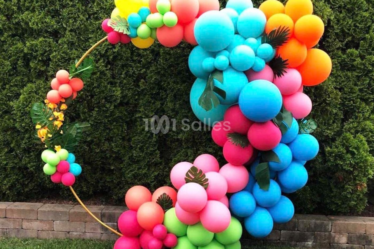 Arc décoratif en ballons colorés avec des feuilles tropicales, placé devant une haie de verdure.