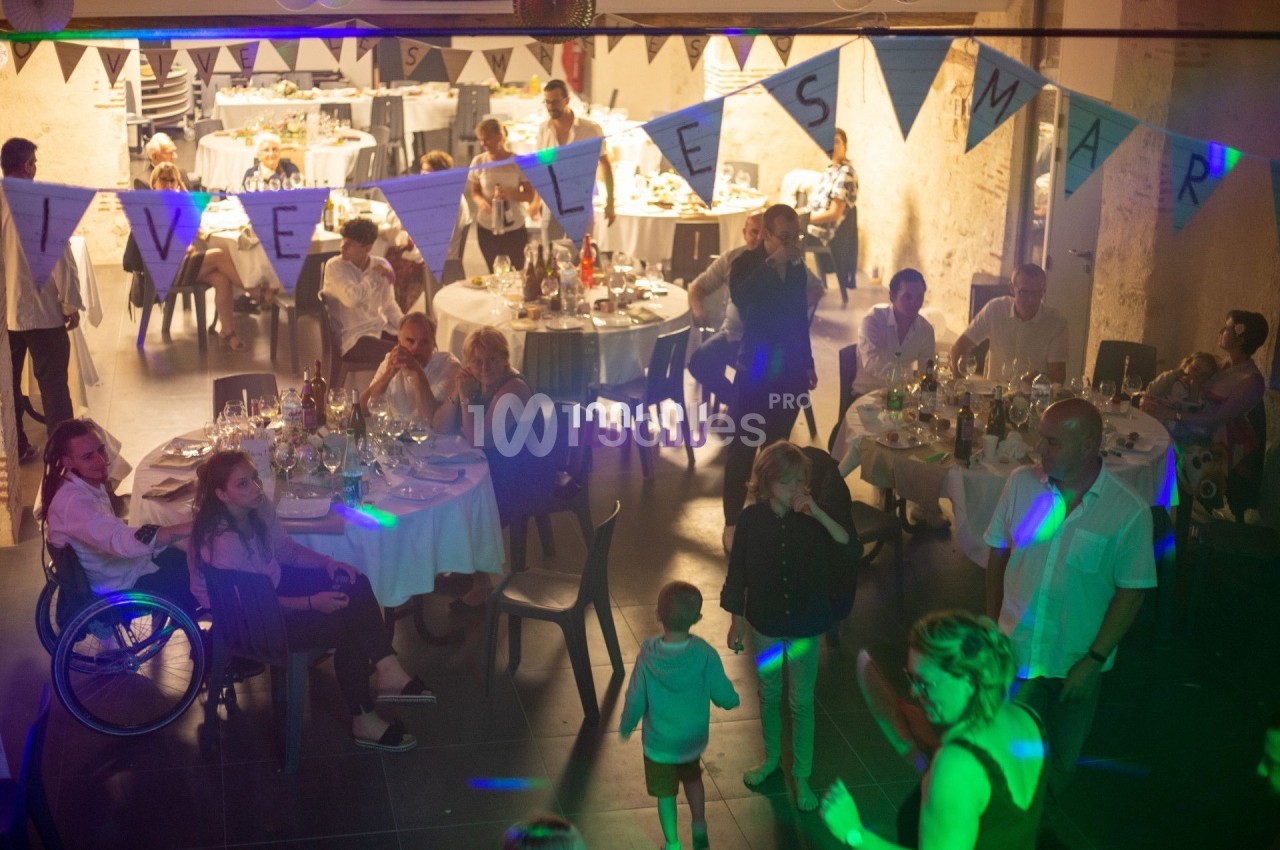 Des invités assis et debout dans une salle décorée pour une fête, avec une bannière ’Vive les mariés’.