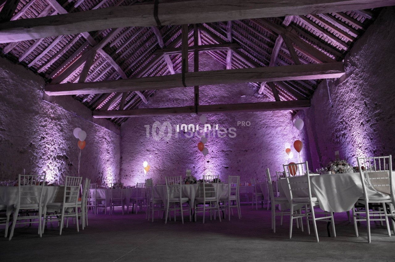 Salle en pierre avec charpente apparente, éclairée en violet, décorée de ballons et tables dressées pour un événement.