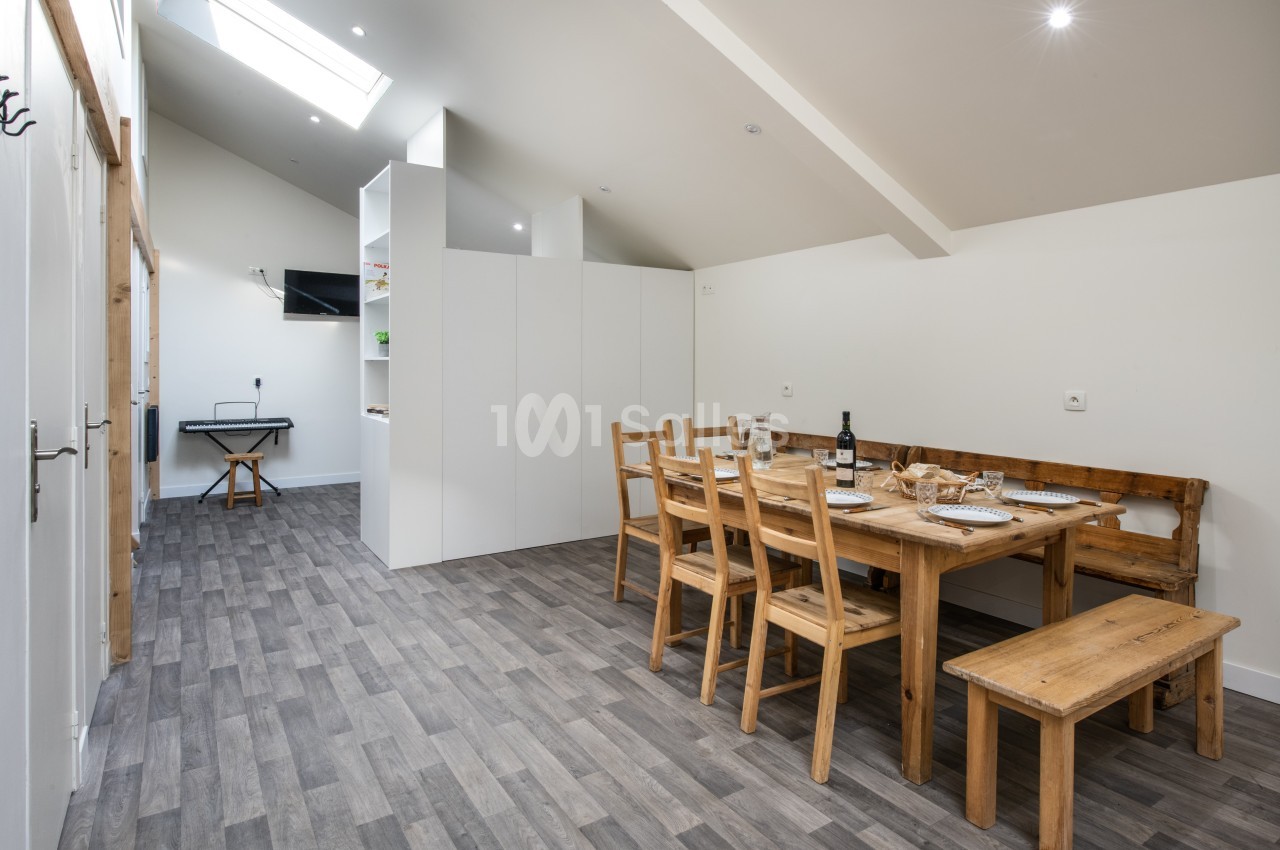 Salle à manger lumineuse avec table en bois, chaises assorties, sol en parquet gris et cuisine ouverte en arrière-plan.