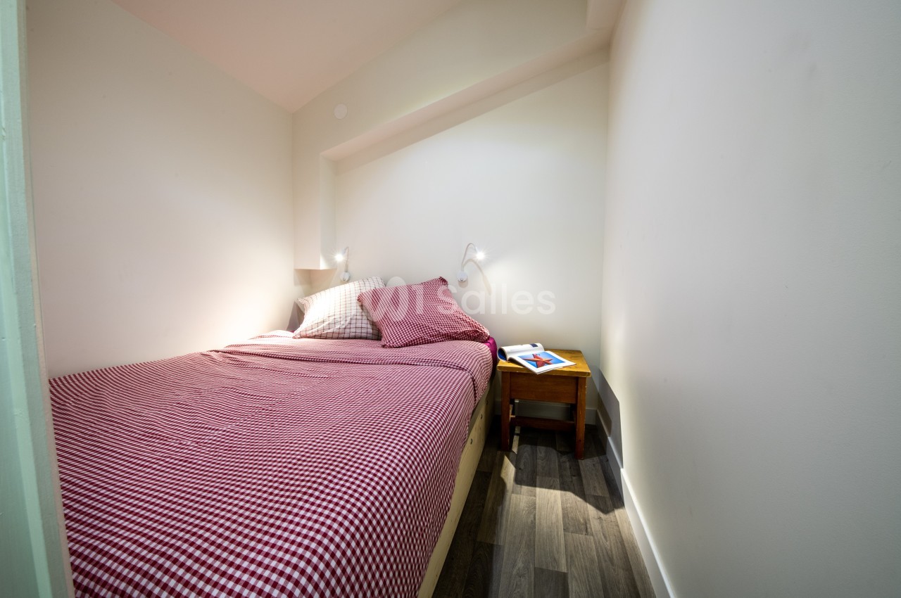Lit double avec draps à carreaux rouges dans une petite chambre lumineuse, table de chevet avec lampe et livre.