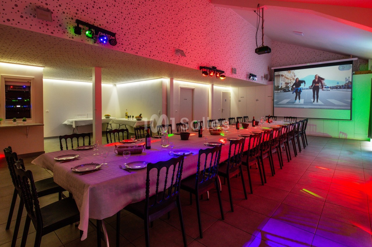 Salle de réception avec une longue table dressée, éclairages colorés et un écran projetant une vidéo.