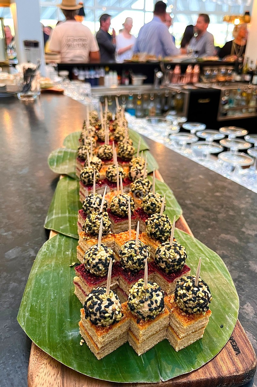 Plateau de bouchées apéritives sur feuilles de bananier, avec des invités en arrière-plan dans un bar animé.
