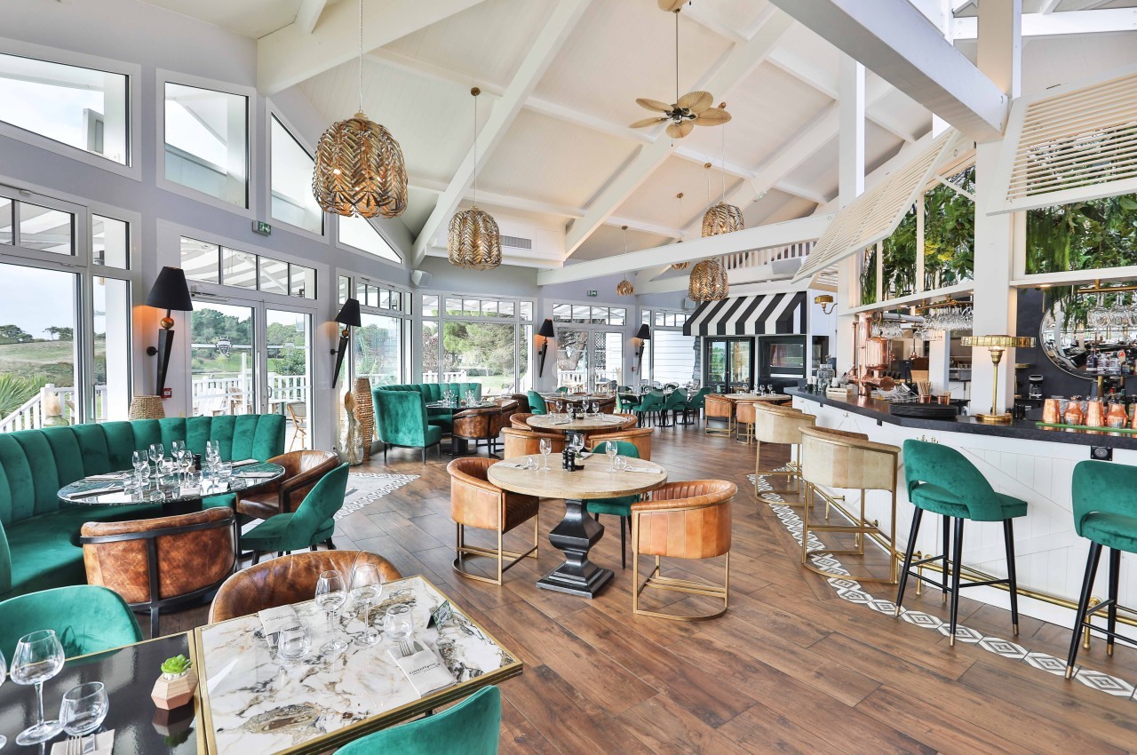 Salle de restaurant lumineuse avec grandes baies vitrées, mobilier élégant et bar central moderne.