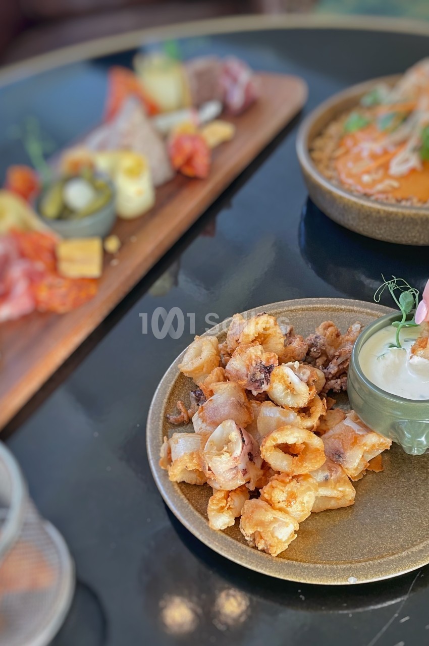 Assiette de calamars frits avec une sauce à tremper, accompagnée de plats variés en arrière-plan sur une table.