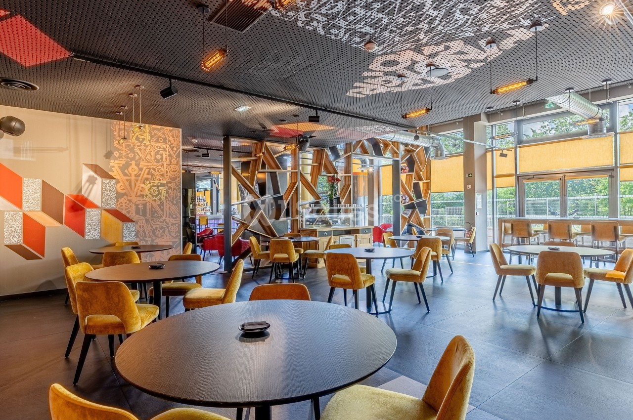 Salle de restaurant moderne avec chaises jaunes, tables rondes noires et grandes baies vitrées laissant entrer la lumière.