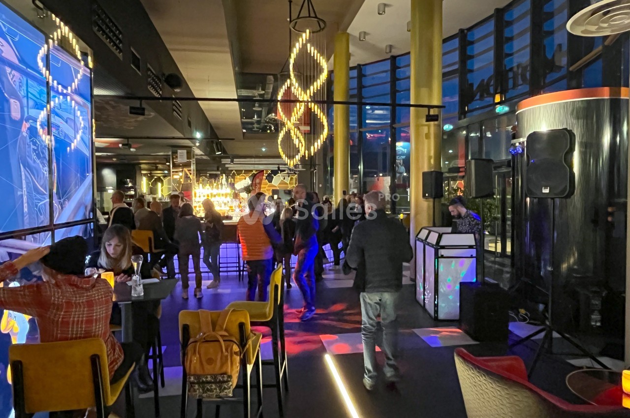 Personnes discutant et dansant dans un bar moderne avec éclairage coloré et grandes baies vitrées en soirée.