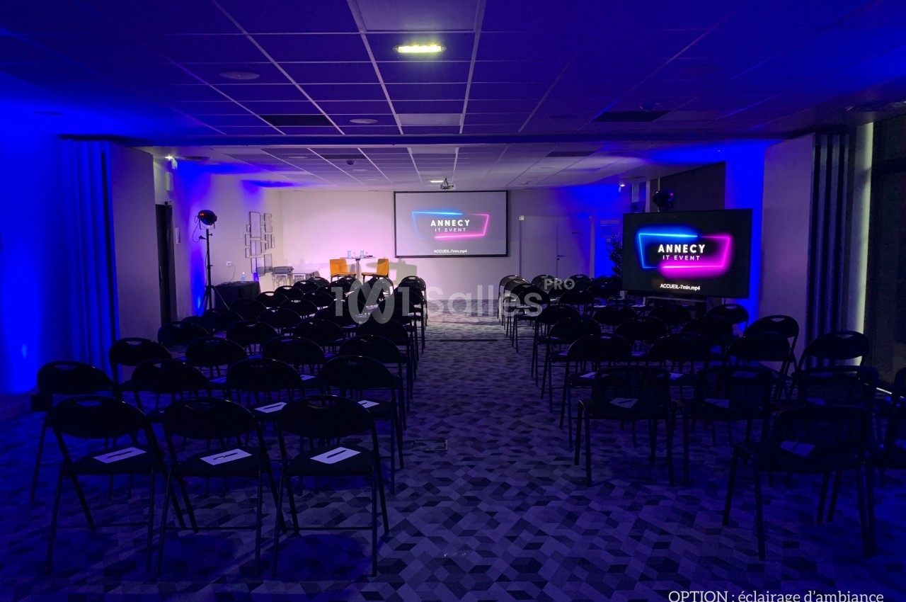 Salle de conférence avec rangées de chaises, éclairage d'ambiance bleu et écran affichant ’Annecy Event’.