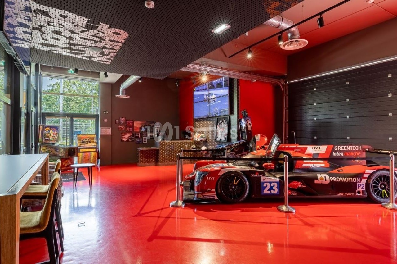 Voiture de course rouge exposée dans une salle moderne avec sol rouge, mobilier en bois et baies vitrées.