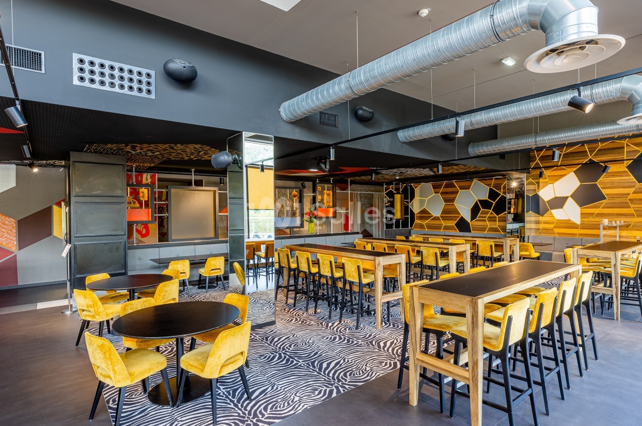 Salle moderne avec tables hautes et basses, chaises jaunes, décor géométrique coloré et conduits apparents au plafond.