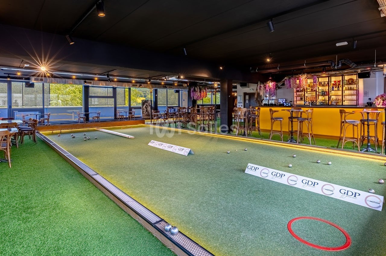 Terrain de pétanque intérieur avec sol en gazon synthétique, entouré de tables et d'un bar éclairé en arrière-plan.