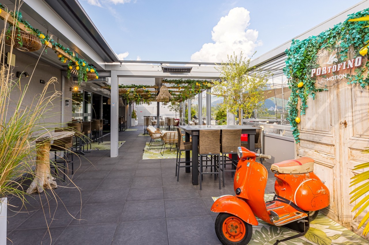 Terrasse aménagée avec tables, chaises, décoration végétale et un scooter orange sous un ciel dégagé.