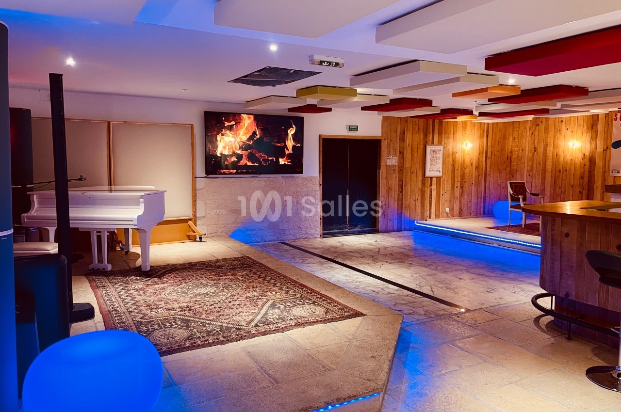 Salle avec un piano blanc, un tapis, un écran affichant un feu de cheminée, et un éclairage chaleureux.
