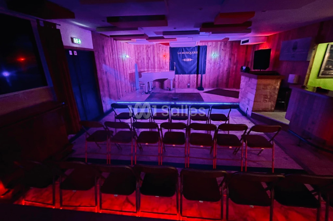 Salle de spectacle avec scène éclairée, piano blanc et rangées de chaises vides dans une ambiance tamisée.