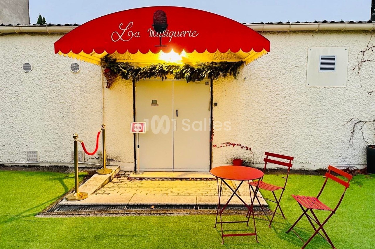 Entrée d'un bâtiment avec une enseigne rouge ’La musiquerie’, une table et des chaises rouges sur une pelouse artificielle.