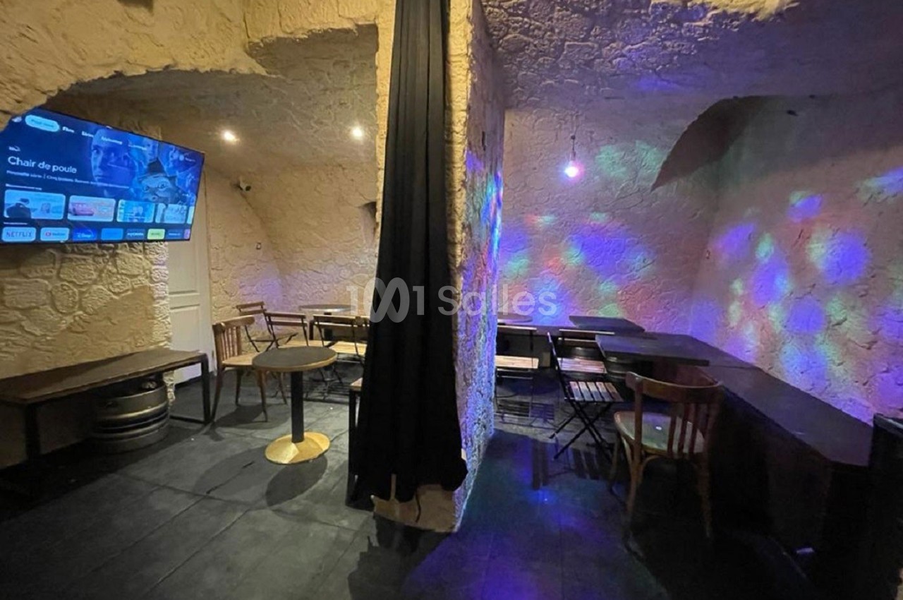Salle voûtée avec murs en pierre, tables et chaises en bois, éclairage tamisé avec projections lumineuses colorées.