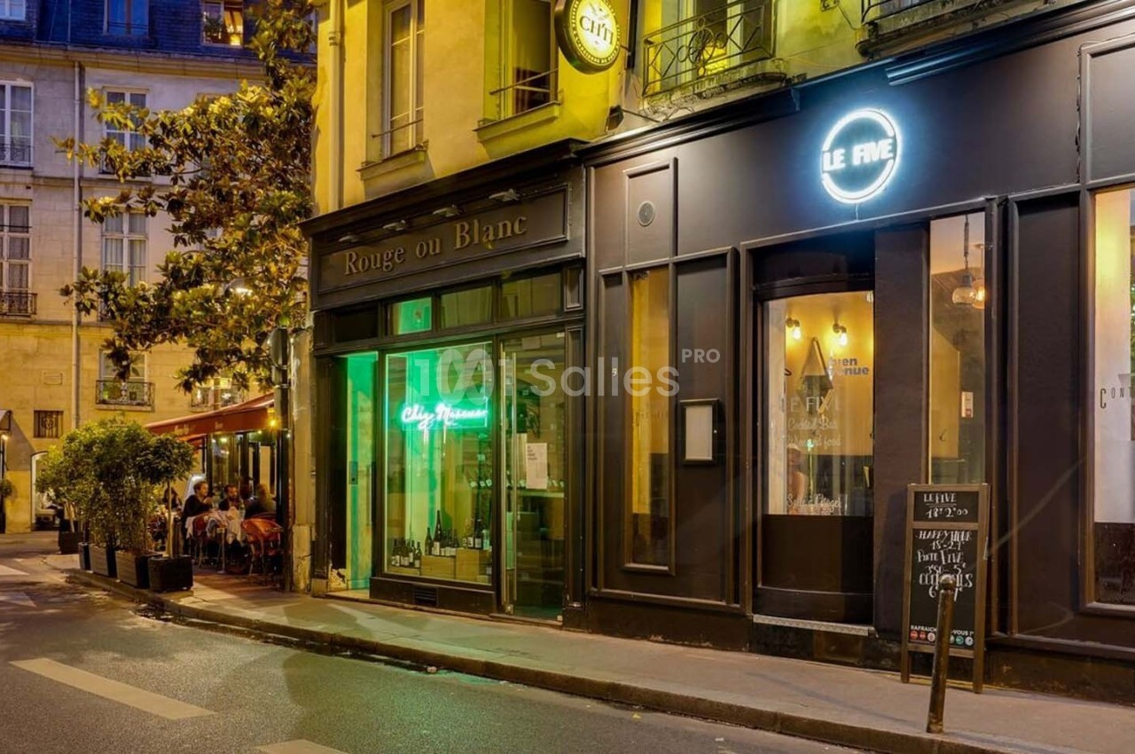 Façade de restaurants et bars éclairés en soirée dans une rue calme avec quelques tables en terrasse.