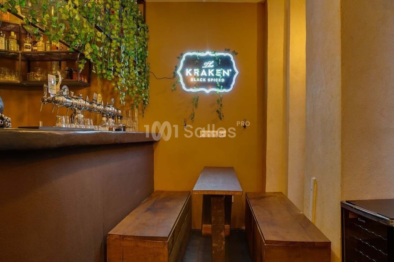 Bar avec comptoir en bois, étagères de bouteilles, plantes suspendues et enseigne lumineuse murale.